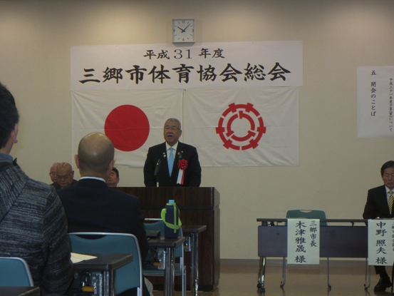 「平成31年度三郷市体育協会定期総会」の紙が貼られている部屋で、パイプ椅子に関係者たちが着席しているなか、演壇に立つ市長が挨拶をしている写真