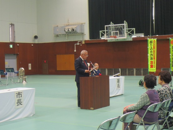 床にグリーンのシートが敷かれている体育館で、パイプ椅子に座っている関係者たちの前に演壇が設置され、市長が話をしている写真