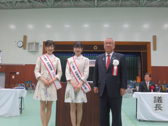 グリーンのシートが敷かれた体育館で、胸章を着けた市長が三郷市PR大使のMika+Rikaさんと一緒に並んで記念撮影をしている写真