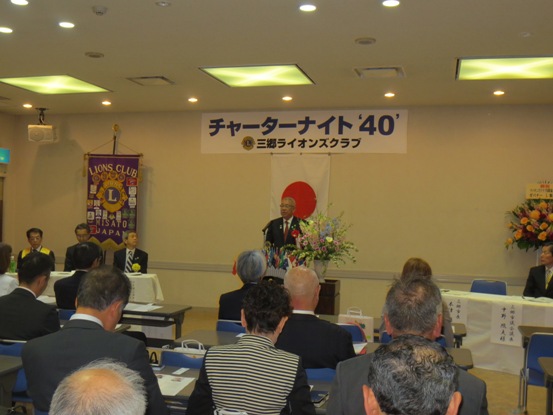 壁に「チャーターナイト40」と書かれた紙が貼られ、部屋の左側にはライオンズクラブの旗が設置されている部屋で、多くの関係者が着席しているなか、演壇に立つ市長が挨拶をしている写真