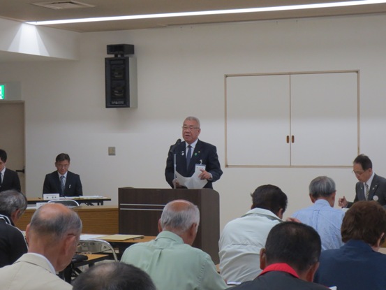 作業着やスーツ姿の関係者たちが椅子に着席している部屋で、演壇に立つ市長が資料を持って話をしている写真