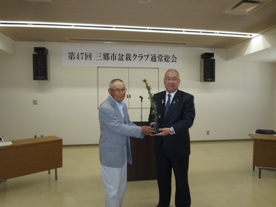 「第47回三郷市盆栽クラブ通常総会」の紙が貼られた部屋で、市長とグレーのスーツ姿の男性が一緒に苗木を持って記念撮影をしている写真