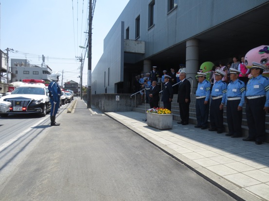 会場外の広場で警察官の制服を着た関係者たちと一緒に並んで立っている市長と、道路に停められたパトカーの近くに立って市長たちと向かい合わせになっているヘルメット姿の関係者の写真