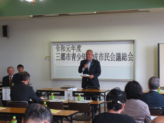 ホワイトボートに「令和元年度三郷市青少年育成市民会議総会」と書かれた紙が貼られ、長机が等間隔に設置されている部屋の正面に立って話をしている市長の写真