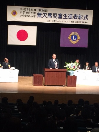 ステージ上部に「無欠席児童生徒表彰式」と書かれた横断幕が設置され、演台に立つ三郷市長がお祝いの挨拶の言葉を述べている写真