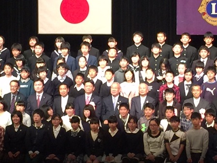 三郷市長が受賞した子どもたちと関係者とともに並び、国旗とライオンズクラブの旗を背景に記念撮影をしている写真