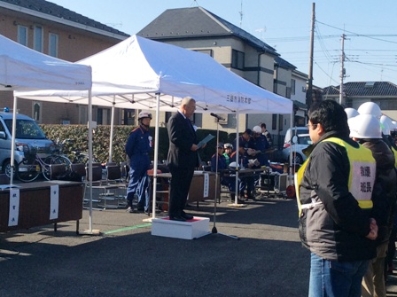 テントの前に設置された小さな台の上に立っている市長の話を向かい合って立っているヘルメットを被った参加者が聞いている様子を左側から撮影した写真