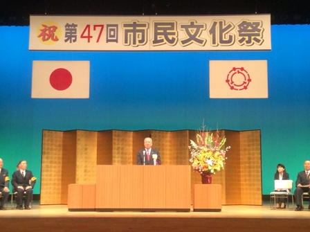 「第47回市民文化祭」と書かれた横断幕や国旗と三郷市旗が飾られたステージの金屏風の前の檀上で挨拶を行っている市長の写真