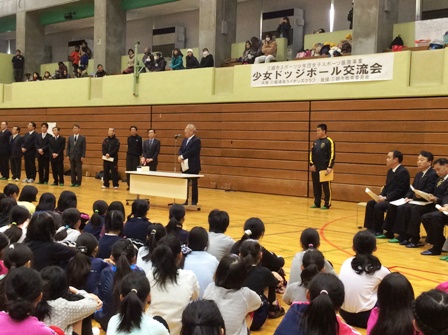 「少女ドッジボール交流会」と書かれた横幕が貼られた体育館のマイクスタンドの前に立つ市長の話を体育座りをして聞いている子供たちの後ろ姿の写真