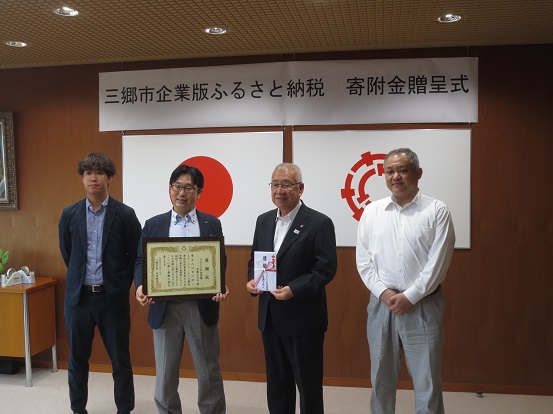 「三郷市企業版ふるさと納税 寄附金贈呈式」と書かれた吊り看板の設置された部屋で表彰状を持った男性と封筒を持った市長を中心に、両側に一名ずつ関係者たちが並んで記念撮影をしている写真