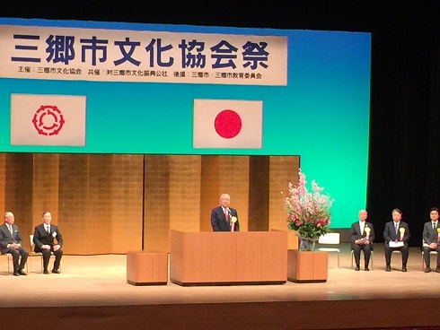 「三郷文化協会祭」と書かれた横断幕と、国旗、三郷市の旗が掲げられた舞台の上で、金の屏風を背に演台に立ち、客席に向かって挨拶をする市長と、演台の両脇に配置された椅子に座る来賓の写真