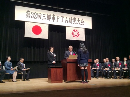 「第32回三郷市PTA研究大会」と書かれた横断幕や国旗と三郷市市旗が飾られたステージの演壇の前に立つ女子高生と向かい合っている市長が表彰状を読み上げている様子の写真