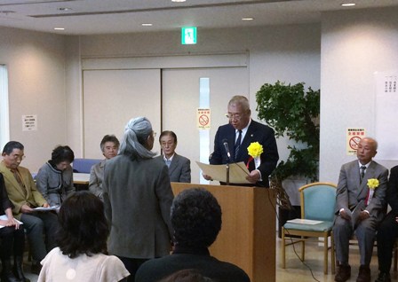 演壇を挟んで男性と向かい合って立っている市長が表彰状を読み上げている様子の写真