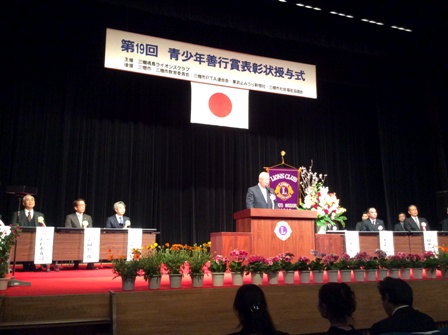 「第19回青少年善行賞表彰状授与式」と書かれた横断幕と国旗が飾られたステージの檀上で挨拶を行っている市長の写真