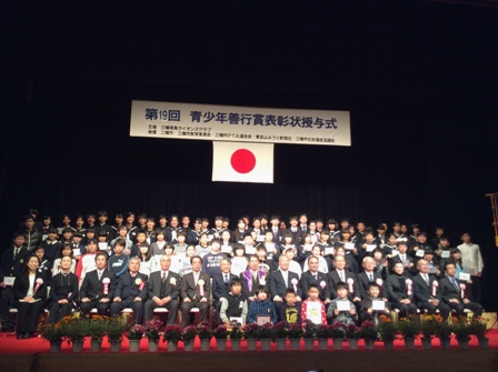 ステージ上で市長を挟んで胸章リボンをつけた方々が前列の椅子に座り、小さな子供たち6名が前に、他の大勢の受賞者が後ろに立っている記念写真