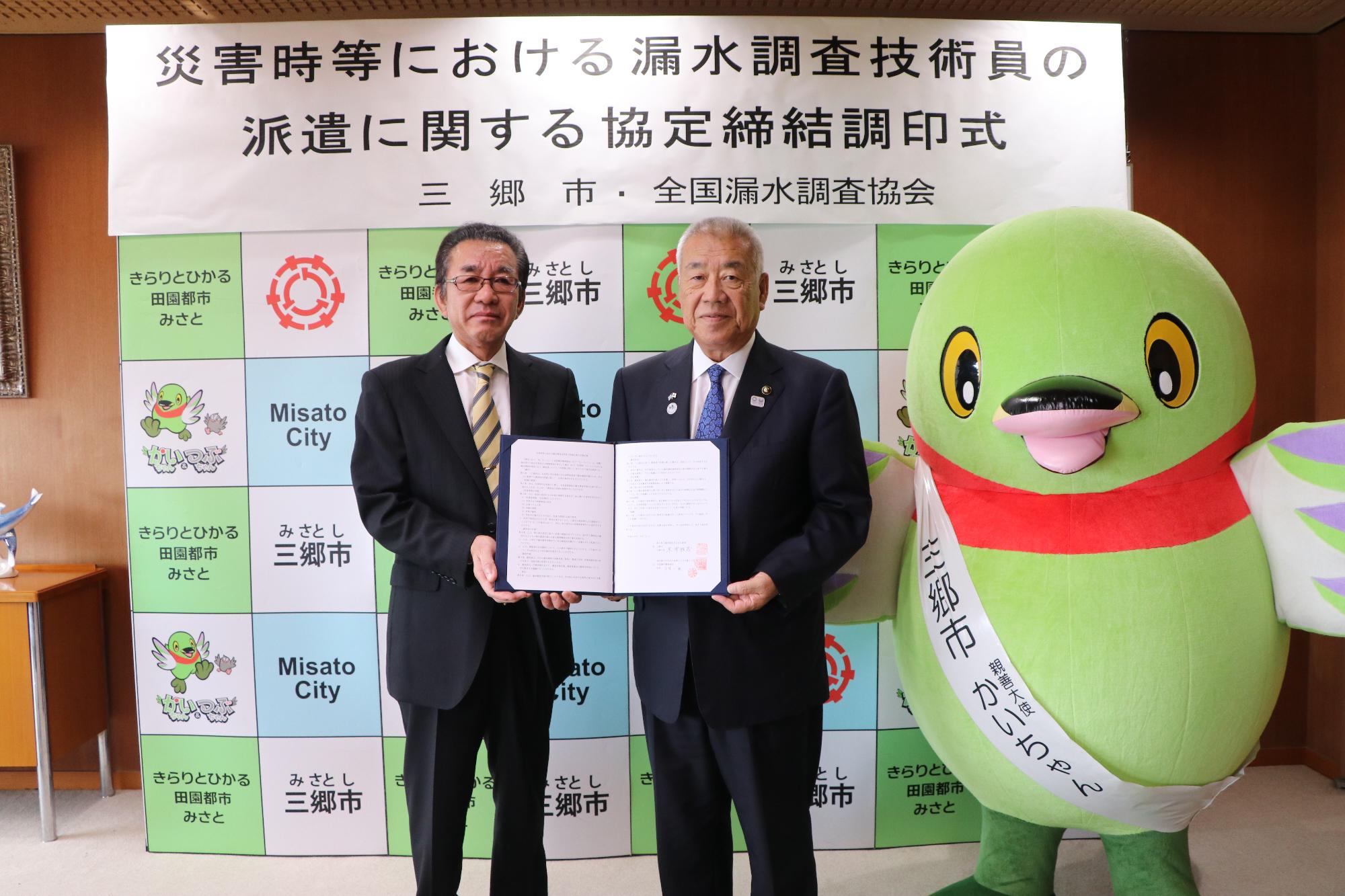 調印書を市長と男性関係者が一緒に持ち、その横に三郷市キャラクターかいちゃんが立っている写真