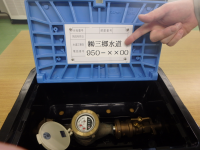 メーターボックスの青色の蓋に貼ってある施工業者の連絡先のプレートを指さし示している写真