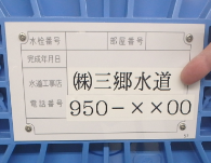 青色の蓋に貼ってある施工業者の連絡先のプレートを指さし示している写真