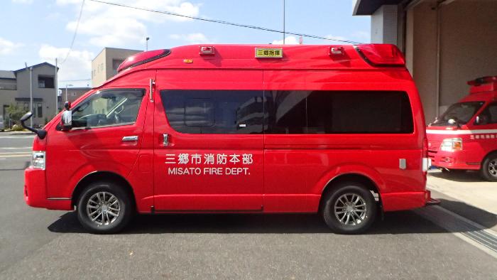 赤い指揮車を左側から写している写真。ボディには白文字で三郷市消防本部と印字されている。