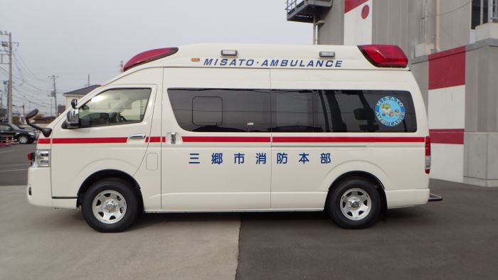引き戸式のフロントドア部分に青文字で三郷市消防本部と記載されていて、後方ドアガラスに三郷市のゆるキャラかいちゃん＆つぶちゃんの丸いシールが貼られた消防車の側面部分を屋外で撮影した写真