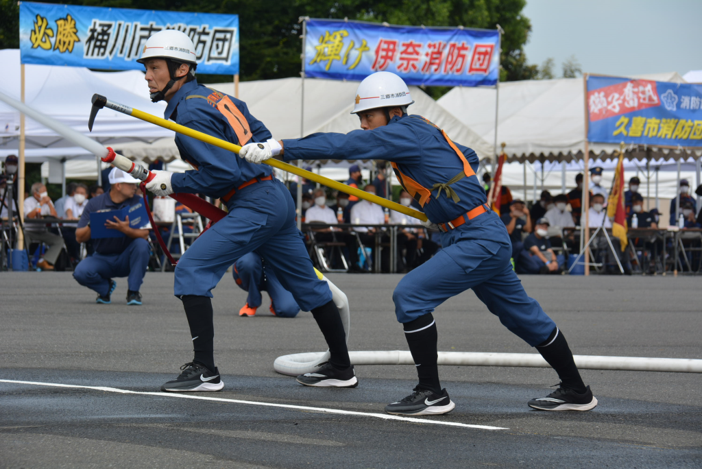 ヘルメットと制服を身につけた男性2人が、消防鳶や消火用ホースを持って構えている様子と、複数張られた白いテントの下でその様子を見物する多くの人々の写真