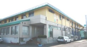 レモン色の外壁にエメラルドグリーンのアクセントがある三郷市立鷹野学校給食センター東館の入口から斜めに外観を写した写真