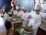 教室の黒板を背に、白い給食着と帽子、マスクを身につけた生徒が4人並んで立ち、前の机に置かれた食缶から、汁物やおかずを器に盛っている様子と、その正面に立って、手に持ったトレーに順番に器を乗せてもらっている生徒たちの写真
