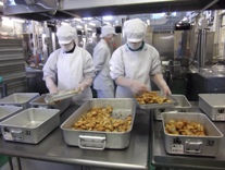 大きなアルミ容器に給食を入れている白衣姿の2名の方とその後ろに立つ1名の白衣姿の作業時の写真