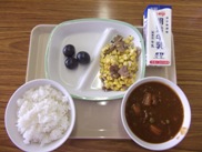 ピンクのトレーに給食が右上から半時計周りで牛乳、コーン、ブドウ、ごはん、カレーと並んでいる写真