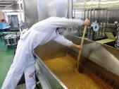 調理室内の、大型の銀色をした煮炊き釜に調理中のカレーのようなものが大量に入っており、それを、白衣と帽子、マスクを身につけた男性が、釜の上に身を乗り出しながら、両手で持った長い棒でかき混ぜている様子の写真