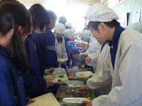 右側に給食当番の人達が並び、その前に給食をとりに来ている生徒たちを横から写した写真