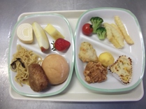 トレーにお皿が2枚並べられ左側のお皿にパン、焼きおにぎり、焼きそば、いちご、リンゴ、パインアップル、ミニクレープロール、右側のお皿にポテト、ブロッコリー、ミニトマト、フリッター、唐揚げ、卵焼き、ミニグラタンが並べられた給食の写真