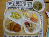 トレーに盛り付けられたパン、焼きおにぎり、おかず、フルーツ、ケーキなどを上から写した給食の写真