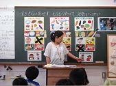 「すききらいをせず、もりもりたべて、元気いっぱい」と書かれた下に、食べ物のイラストなどが印刷された紙がたくさん貼られた黒板を背に、正面を向いて教壇の前に立っている女の先生が、右下のイラストの方向いて、指をさしている様子と、黒板の方を向いて座っている生徒たちの写真