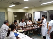 L字型に並べた長机に透明のパックに入った料理がいくつか並べられており、椅子に座った3人の男女が皿に取った料理を食べている様子と、それを見ている、書類を広げた机に向かい座っている白衣の男女と、机の前の空間に立つ白衣の女性たちの写真