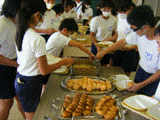 子供たちが各自のトレーを持ち、テーブルの両端から給食を取ろうとしているバイキング給食の様子を写した写真