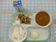 トレーに給食が右上から半時計周りでカレー、副菜、ごはん、ゆで卵、トレー、副菜の後ろに牛乳が並んでいる写真