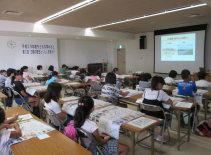 前方のスクリーンに映した出された映像を見ながら学習している子どもたちを後方から写した写真