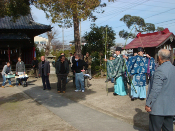 住民が見守る中、装束を身に着けた2名の神主が、境内で弓を射ろうと構えている写真
