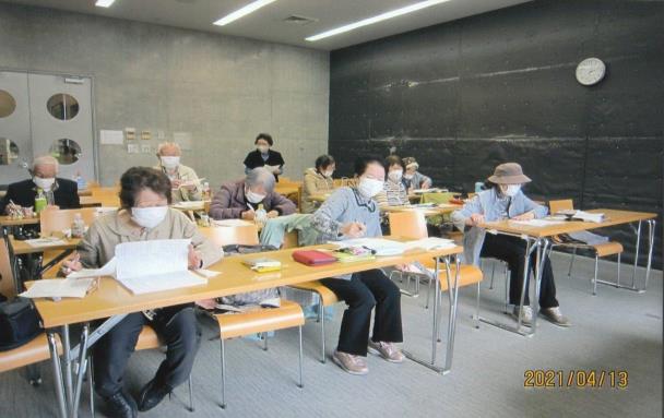 コンクリートの壁の室内に複数の長机と椅子が並べられ、そこにたくさんの高齢の男女が座って、机に置かれた紙に向かっている句会の様子の写真