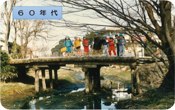 用水路に架かる木製の橋の上に立つ数名の園児がこちらを向いている昭和60年代の三郷市の写真