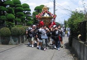 きれいに剪定された植木の横の道路を、神輿を担いで歩く人たちの写真