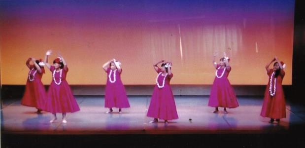 壁が青やオレンジ色のグラデーションになった舞台上で、赤の衣装に白のレイを身に着けた6人の女性が両手を挙げながらフラダンスを披露している写真