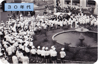 白い服を着て帽子をかぶったたくさんの学生が噴水の周りに集まっている昭和30年代の三郷市の白黒写真