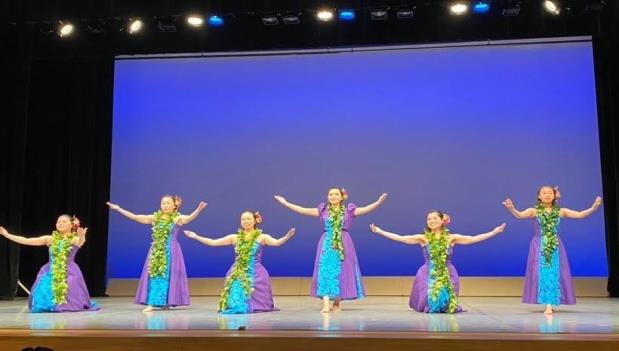 紫と水色の衣装に黄緑の花の首飾りをつけた6名の女性が、ステージ上で両手を広げて踊っている様子の写真