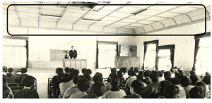 奥の檀上に立っている男性の話を参加者が座って聞いている後ろ姿の会場のカーブを描いた折上げ造りとなっている天井が黒線で記されている白黒写真