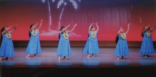 6人の女性が首にレイを付け、水色の衣装に身を包み、両手を斜め上に上げて踊っている写真