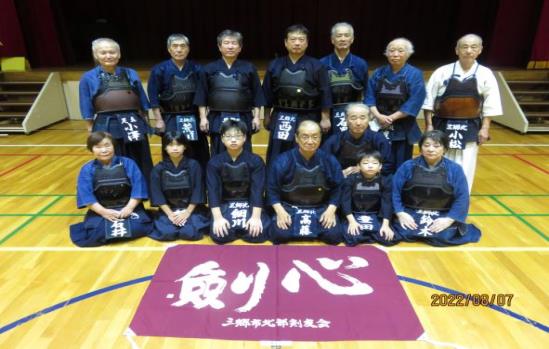 体育館の舞台の前で、剣道着に胴を身につけた子供から年配の男女が、前列は正座、後列は立って2列に並び、その手前の床に大きく「剣心」と書かれた赤紫色の応援幕が広げられている、三郷市北部剣友会全員の集合写真