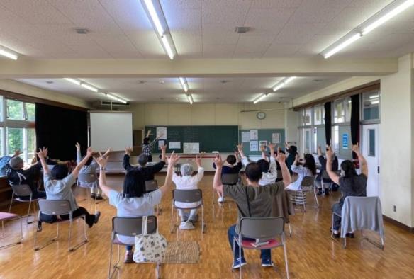 教室の黒板の前に講師の人が立ち、椅子に座っている15名ほどの参加者達が、講師の方と同じように両手を挙げているところを後法から撮った写真