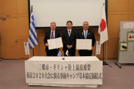 中央に3名の関係者の男性が横一列に並び後方には、両端にギリシャと日本の国旗が置かれ左右の男性が協定書を持って立っている締結式の様子の写真